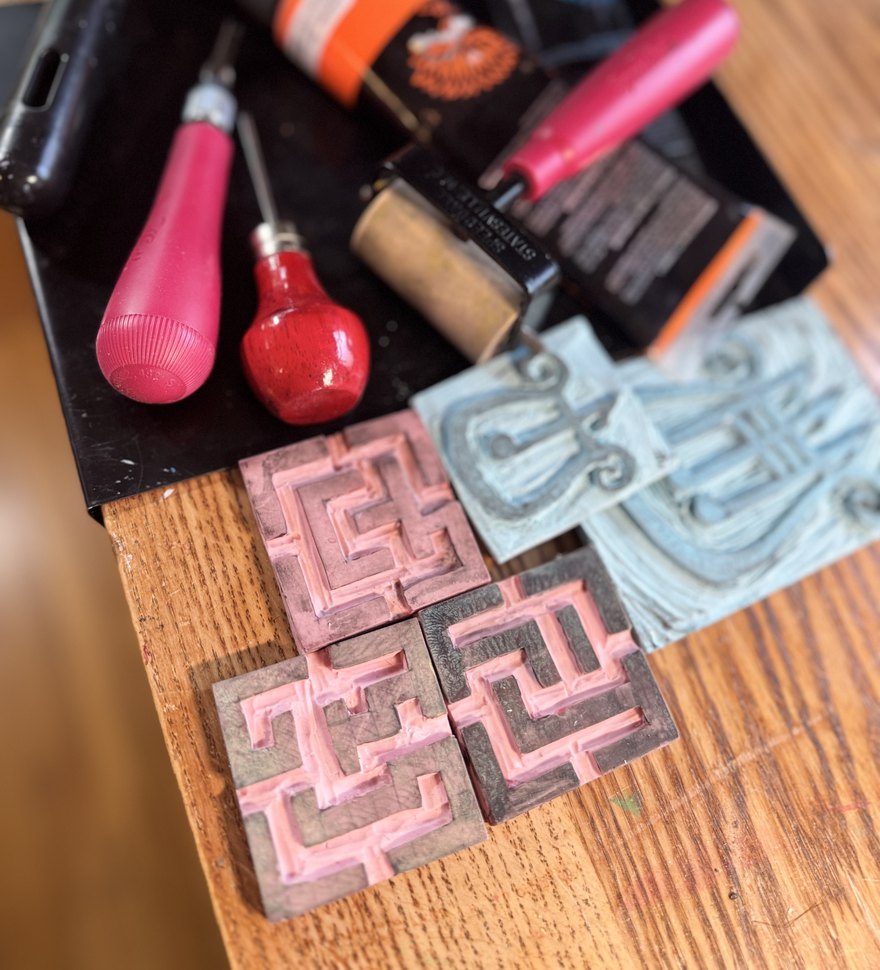 Carving gouges, a brayer and ink on a workbench, beside the carved lino blocks for four maze tiles and the lyre.
